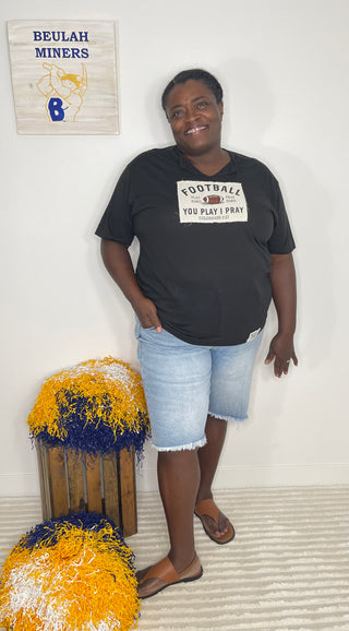 A woman stands indoors wearing a black graphic T-shirt with a white football-themed print that reads 'FOOTBALL YOU PLAY I PRAY' and light blue denim shorts. She is posed in front of a white wall with a 'BEULAH MINERS' sign above and yellow-and-blue decorative pom-poms on a crate below.